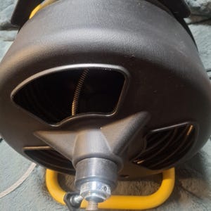 May include: A black plastic drain cleaning machine with a yellow metal frame. The device features a central drum housing a coiled metal cable, with a silver-colored metal tip extending from the front center and a black adjustment knob.