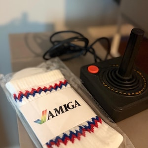 May include: A pair of white socks with a red and blue zigzag pattern at the top, packaged in clear plastic with a white label featuring the Amiga logo. Next to the socks is a black joystick controller with a single red button and a black cable.