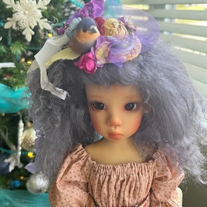Puede incluir: Una muñeca con el pelo rizado gris que lleva un vestido rosa y un sombrero floral con un pájaro azul en la parte superior.