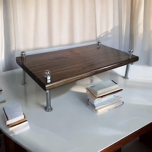 Peut inclure: Un rehausseur de bureau en bois brun foncé avec des pieds métalliques et des accents argentés. Le rehausseur est posé sur un bureau blanc avec une pile de livres en dessous.