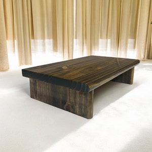 Puede incluir: Una plataforma de madera marrón oscuro con un borde ligeramente redondeado. La plataforma se eleva sobre dos patas cortas.