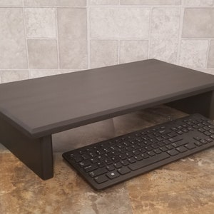 May include: A dark brown wooden monitor stand with a keyboard underneath. The stand is raised off the ground and has a shelf for the keyboard.