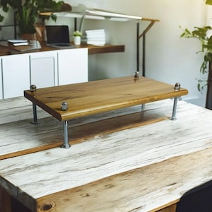 Puede incluir: Un soporte para portátil de madera con patas de metal se encuentra sobre una mesa de madera blanqueada. El soporte está hecho de madera natural con un acabado marrón claro. Las patas son de metal plateado y tienen un diseño sencillo.