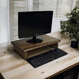 May include: A dark brown wooden monitor stand with a black computer monitor and keyboard on a light wood desk. A small green plant is in the background. The stand is 12 inches wide.
