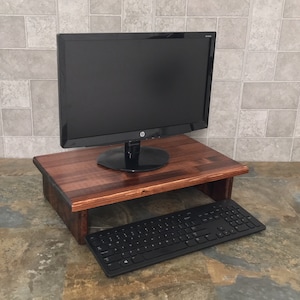 Puede incluir: Un soporte de monitor de madera marrón oscuro con un monitor de computadora y un teclado negros. El soporte es rectangular con un estante inferior para el teclado. El monitor es negro con una base circular.