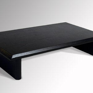 May include: A black wooden riser with a rectangular top and a cutout in the front. The riser is about 12 inches long and 6 inches wide.