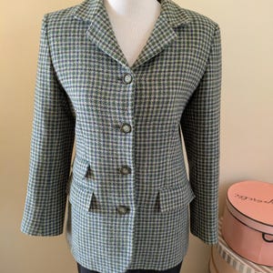 Puede incluir: Una blazer de pata de gallo verde, azul y beige con tres botones y dos bolsillos delanteros. La blazer se exhibe en un maniquí y tiene un diseño clásico y entallado. La chaqueta es de peso medio y adecuada para climas más fríos.