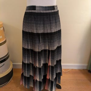 Puede incluir: Una falda larga plisada con un estampado de cuadros grises, negros y rosas. La falda se exhibe en un maniquí, mostrando toda su longitud y diseño. La tela parece ser un material tejido, con un estilo clásico y atemporal.