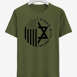 May include: An olive green t-shirt with a circular design incorporating the American flag and a Star of David. The black text reads "I STAND WITH ISRAEL". The t-shirt is hanging on a white hanger.