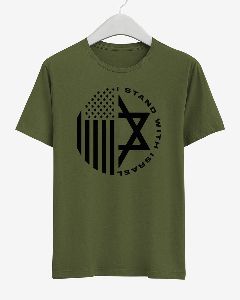 May include: An olive green t-shirt with a circular design incorporating the American flag and a Star of David. The black text reads "I STAND WITH ISRAEL". The t-shirt is hanging on a white hanger.