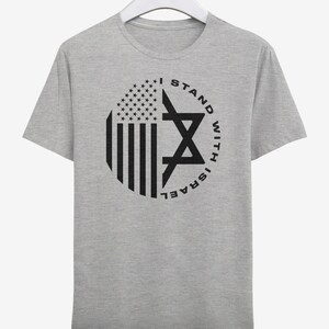May include: A grey t-shirt with a circular graphic incorporating the American flag and a Star of David. The black text reads "I STAND WITH ISRAEL". The t-shirt is displayed on a white hanger.