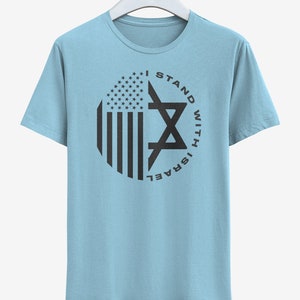 May include: Pale blue t-shirt with a circular design incorporating the American flag and a Star of David. The black text reads "I STAND WITH ISRAEL". The t-shirt is displayed on a white hanger.
