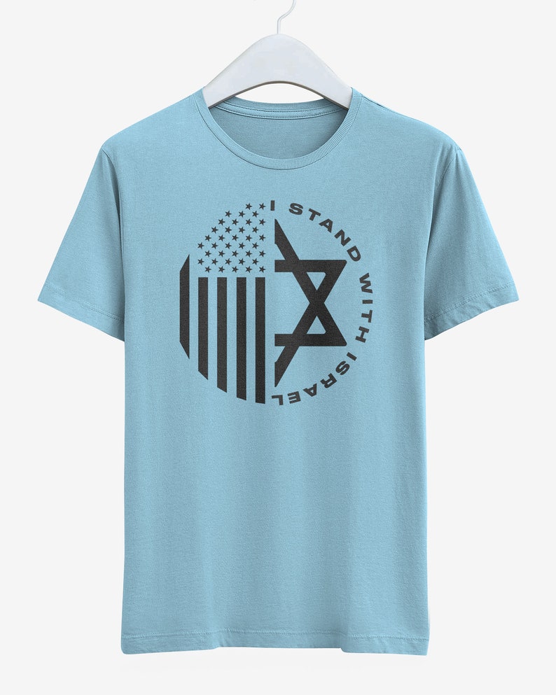 May include: Pale blue t-shirt with a circular design incorporating the American flag and a Star of David. The black text reads "I STAND WITH ISRAEL". The t-shirt is displayed on a white hanger.