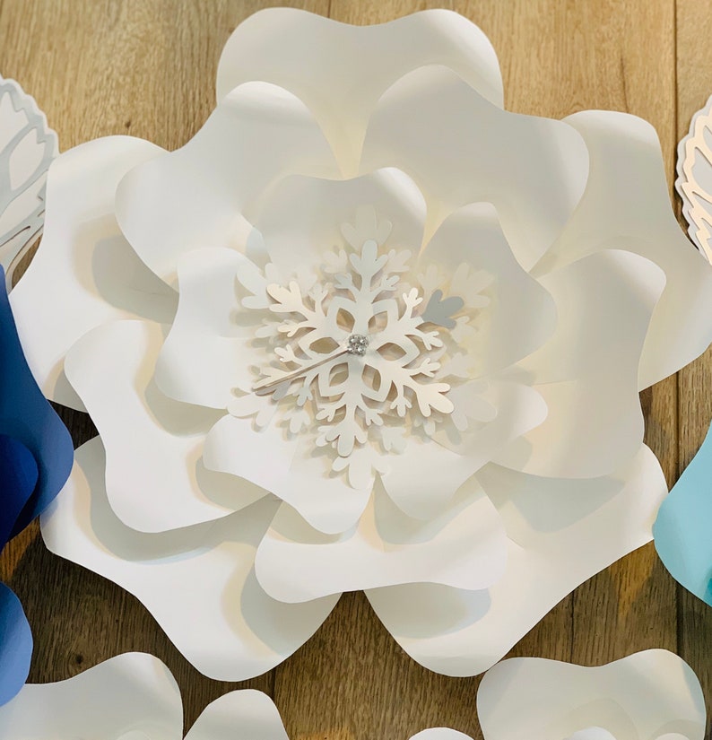 May include: A large white paper flower with a snowflake in the center. The snowflake is white with a silver glitter center.