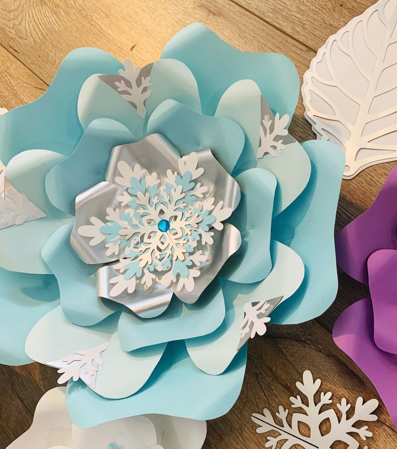 May include: A large, light blue paper flower with a silver and white snowflake in the center. The flower is made of multiple layers of paper and has a textured, ruffled appearance.