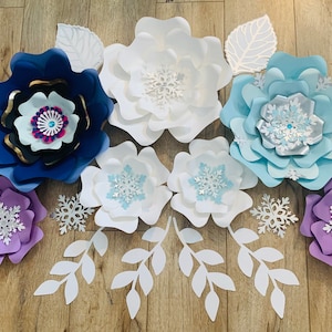 May include: A set of paper flowers in shades of blue, purple, and white, with snowflake designs. The flowers are arranged on a wooden surface, with leaves and branches surrounding them.