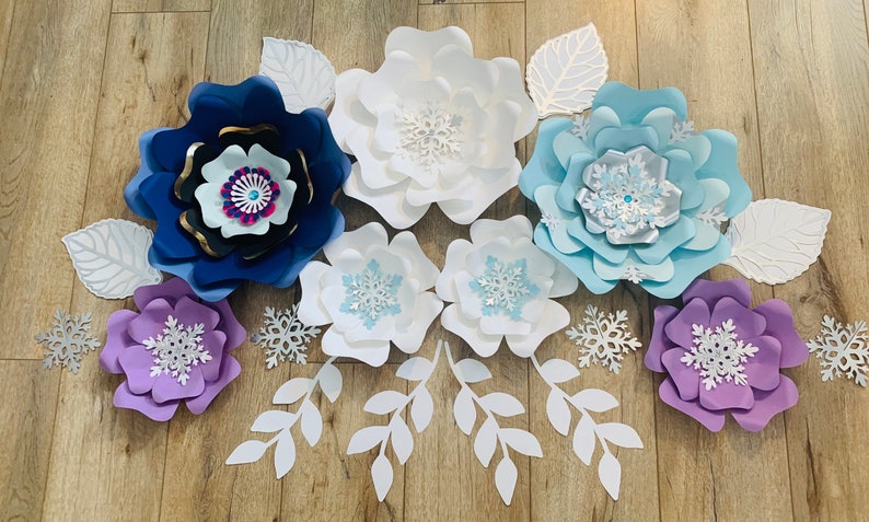 May include: A set of paper flowers in shades of blue, purple, and white, with snowflake designs. The flowers are arranged on a wooden surface, with leaves and branches surrounding them.