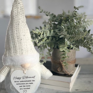 May include: White gnome figurine with a heart-shaped sign that reads 'Happy Retirement Never underestimate the difference you made and the lives you touched.' The gnome is wearing a white knitted hat with silver glitter and has a white beard.