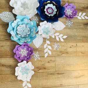 May include: A set of paper flowers in various shades of blue, purple, and white, with snowflake accents. The flowers are arranged on a wood background, creating a festive and whimsical display.