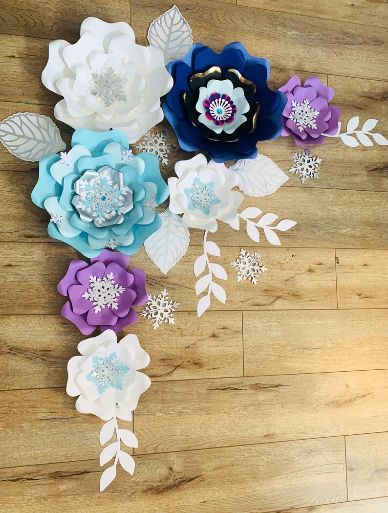 May include: A set of paper flowers in various shades of blue, purple, and white, with snowflake accents. The flowers are arranged on a wood background, creating a festive and whimsical display.