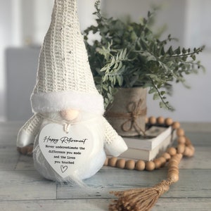 May include: White gnome figurine with a heart-shaped sign that reads 'Happy Retirement Never underestimate the difference you made and the lives you touched.' The gnome is wearing a white crocheted hat with sequins and a white fur trim.