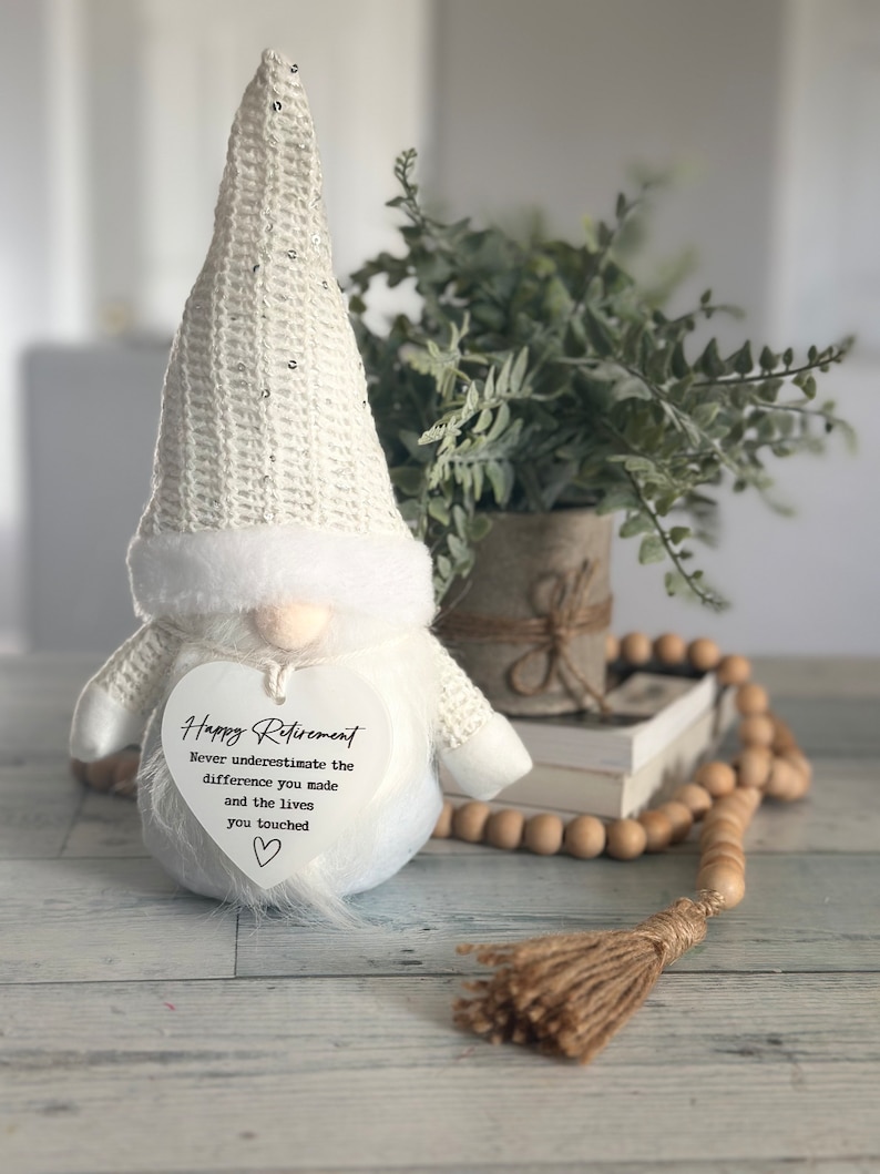 May include: White gnome figurine with a heart-shaped sign that reads 'Happy Retirement Never underestimate the difference you made and the lives you touched.' The gnome is wearing a white crocheted hat with sequins and a white fur trim.