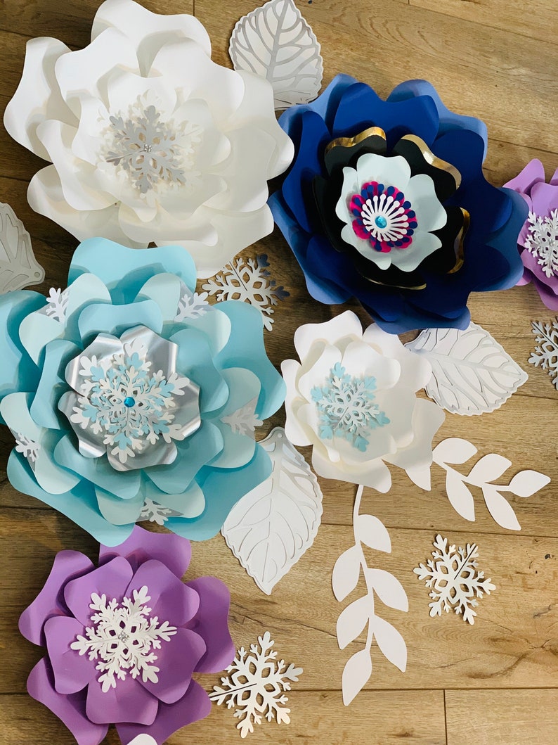 May include: A set of paper flowers in various colors, including white, blue, purple, and black. The flowers are decorated with snowflake cutouts and are arranged on a wooden surface.