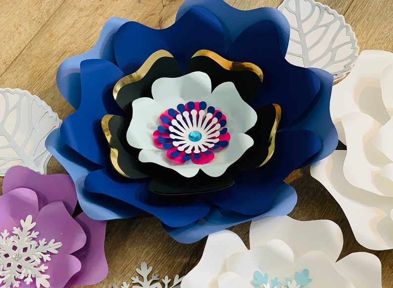 May include: A set of paper flowers in various shades of blue, purple, and white. The flowers are layered and have a 3D effect. The center of the blue flower has a pink and blue design with a sparkly blue jewel.