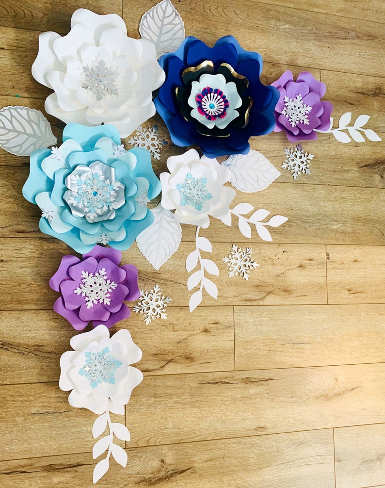 May include: A set of paper flowers in various colors, including white, blue, purple, and gold. The flowers are layered and have a 3D effect. There are also paper leaves and snowflakes scattered around the flowers.