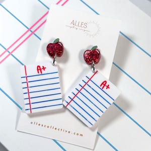 May include: A pair of dangle earrings featuring a red glitter apple charm attached to a white rectangular charm designed to look like a piece of lined notebook paper with a red margin line and the handwritten text A+ in red ink on the top right.