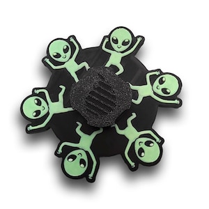 May include: A black phone grip featuring six green alien figures arranged around a central black textured grip. The aliens have large eyes and are in various poses. The background is white.