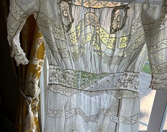 May include: A white sheer dress featuring intricate lace inserts, horizontal ruffled tiers, and three-quarter length sleeves. The bodice includes a wide lace waistband and decorative lace appliques on the chest, all crafted from lightweight fabric.