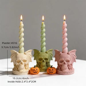 May include: Three skull-shaped candle holders in cream, green, and pink, each featuring bat wings and roses. Each holds a spiral taper candle. Two small orange jack-o'-lanterns sit in front. Dimensions are 10.2 cm or 4 inches wide, 9.7 cm or 3.82 inches high.