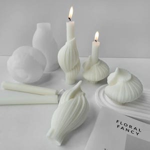 May include: A collection of white, unscented candles in various shapes, including a seashell, a bulbous form, and a stylized flower. Two candles are lit, casting a warm glow. A book with the title "FLORAL FANCY" is visible.