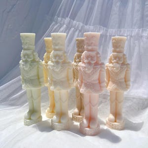 May include: Five decorative nutcracker figurines in various shades of white and pink. The figurines are standing on a white surface.