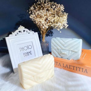 Puede incluir: Un marco de fotos blanco con el texto "PHOTO FRAME" está junto a un ramo de flores secas en un jarrón de cristal. También hay dos jabones decorativos con forma de bolso, uno color crema y otro azul pálido. Los jabones están sobre una superficie blanca.