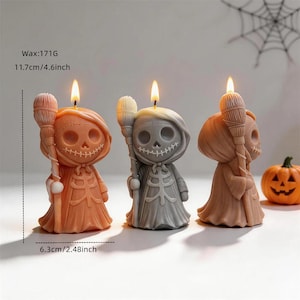 May include: Three skull-shaped candles with lit wicks, each holding a broomstick. The candles are orange, gray, and tan, measuring 4.6 inches tall and 2.48 inches wide, with 171 grams of wax. A small orange jack-o'-lantern sits to the right.