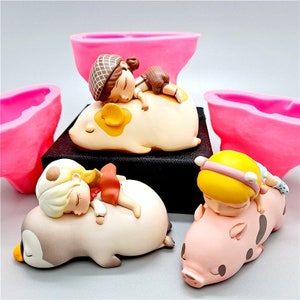 May include: Three figurines of sleeping children atop animal-shaped figures. One child rests on a guinea pig, another on a penguin, and the third on a pig. Pink molds are in the background.