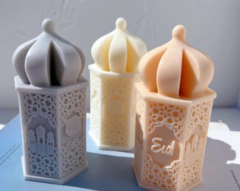 May include: Three decorative, pastel-coloured lantern-shaped objects. Each has a detailed, openwork design on the sides and a domed top. The lanterns are in shades of grey, cream, and peach. The peach lantern has the word "Eid" on it.