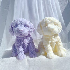 Puede incluir: Dos pequeñas figuritas de perro realistas hechas de cera perfumada. El perro de la izquierda es de color morado lavanda y el perro de la derecha es blanco.