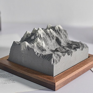 Puede incluir: Una escultura de una cordillera de montañas de hormigón gris en un marco de madera. La escultura está detallada con picos y valles, creando un efecto realista de cordillera.