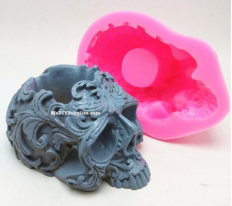 May include: A gray skull-shaped planter with ornate floral detailing, next to a bright pink silicone mold in the shape of a skull. The planter has the text MsDIYSUPPLIES.com on the side.