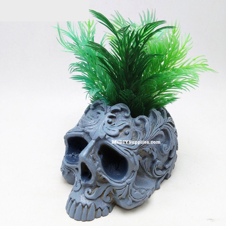 May include: A gray skull-shaped planter with intricate floral carvings, holding vibrant green artificial plants. The planter has a weathered appearance and the text "MDlYSupplies.com" is visible on the side. A unique decorative item.