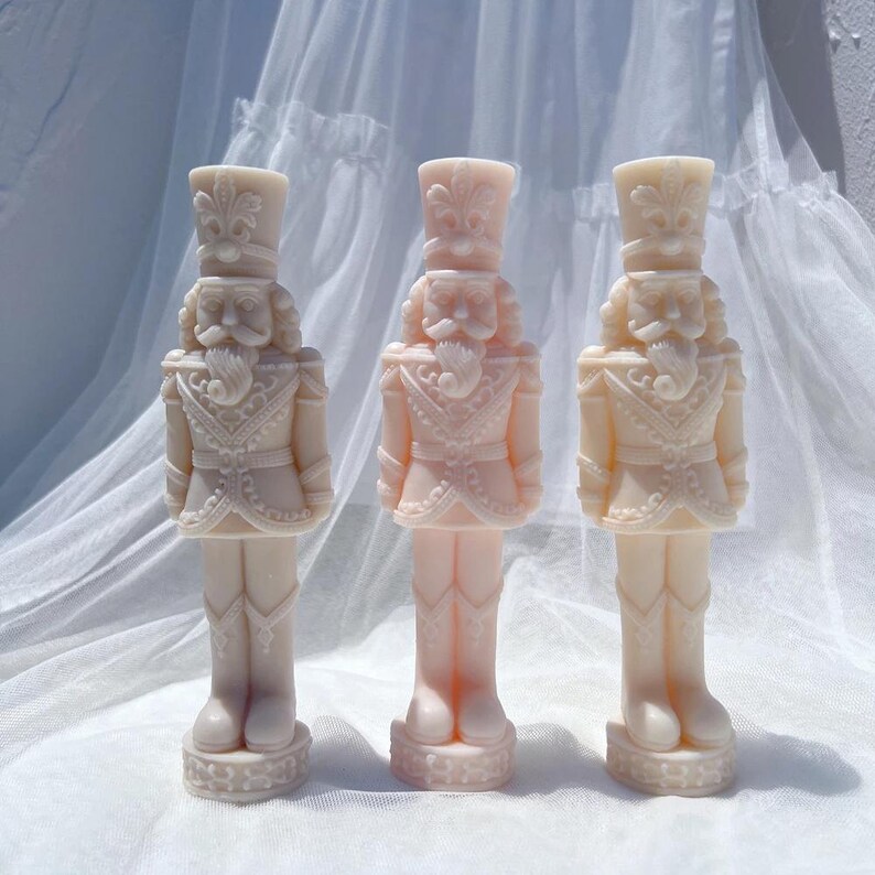 May include: Three white porcelain nutcracker figurines with intricate details. Each figurine is standing tall with a traditional nutcracker design, featuring a hat, mustache, and a detailed uniform.