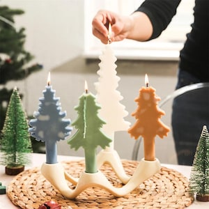 Puede incluir: Un portavelas de cerámica blanca con forma de línea ondulada con cuatro ranuras para velas. Cuatro velas con forma de árbol de Navidad están en las ranuras. Las velas son blancas, azul claro, verde claro y naranja.