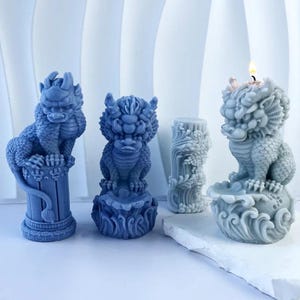 May include: Four blue dragon-shaped candles. Two dragons sit atop decorative bases, one on a pillar, the other on a wave-like design. A third candle is a cylindrical shape with floral carvings. The fourth candle is lit.