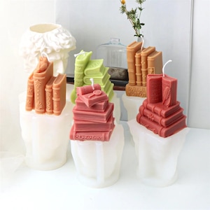 May include: A selection of book-shaped candles in shades of red, green, and brown. The candles are displayed on a white surface, with a decorative bust in the background. Each candle has a wick.