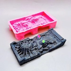 May include: A pink silicone mould with a detailed design of an alien creature. The mould is used to create a black resin casting of the alien creature. The casting is detailed and shows the alien's face, body, and tentacles.