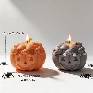 Puede incluir: Dos velas con forma de calabaza, una naranja y otra gris, con caras de calabaza talladas y pequeñas figuras de fantasmas. Las dimensiones son 8,5 cm de alto y 9,2 cm de ancho, con un peso de cera de 303 g. Hay pequeñas arañas alrededor.