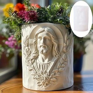 Puede incluir: Una maceta cilíndrica de color crema con un relieve detallado de Jesucristo. La maceta presenta un halo, barba y detalles florales. La maceta está llena de una variedad de flores y vegetación coloridas.